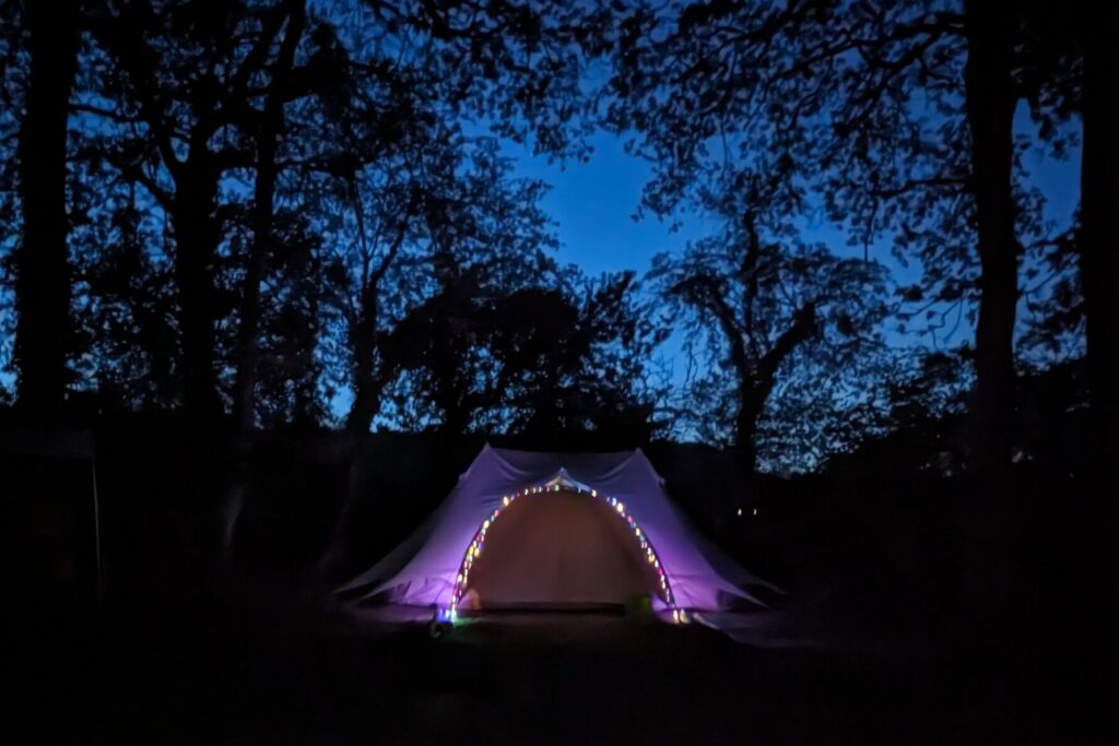 Your beautifully lit Fox Wood Luxury Glamping Bell Tent is super popular. Book now to get your chosen dates.
Glamping Campsite Sussex - South Downs glamping & camping.
Glamping at breathtaking Sussex campsite. Relax in a Luxury pre-pitched Camping tent.  Enjoy Glamping and Sussex camping at South Downs camping forest.
West Sussex glamping in breath taking forest location. Relax amongst trees with a cosy campfire, perfect for creating treasured memories with loved ones.
Our Glamped Emperor Bell Tents are privately located in there own glades, and are great for 4 person families and couples. 
With a bit of extra space for a single family joining in a Woodland Tent Pitch - needs to be booked separately.
What you get when you book a Luxury Emperor Bell Tent Glade?
Your bell tent is equipped with carpets and a comfy bed. 
No inflatable mattress.
There is a double bed and 2 single beds in the tent.
All beds have fresh linen mattress covers, but you do need to pack your own bedding and pillows.
Inside there are magical lanterns for when the sun goes down.
With a remote for your inside chandelier lights, so you can stay snuggled in bed.
The outside lights are solar and light up automatically as it gets dark.
Outside is a campfire area with campfire chairs and log ring tables.
The Fire pit has a grill to cook on too.
And to the side away from the fire is a beautiful farmhouse table with a bench and seats for eating meals and playing cards.
There is a gazebo over the large table just in case of damp weather.
We ask that no food is put inside the tent, so there is a large food cupboard near your dining table to keep all your edibles.
There is no bedding in the bell tent, so you will need to pack duvets and pillows.
You'll find the shared washing and shower facilities nearby.
You'll need to pack:
Bedding, blankets, pillows
Cooking equipment, including pots, pans, utensils
Glasses, crockery and cutlery
Towels and torches
Food and drinks.
But we've taken care of a tent, mattresses, table and seats :-).
There is a large area also in this glade which is suitable for friends to book a separate Woodland Pitch (only 1 extra tent) and they can link it with your booking.  They'll need to book a Woodland Pitch and bring their own tent.
If you are planning to glamp and you are joining friends with tents (more than 1) then you must book the Glamping Bell Tent (suitable for group gatherings) as this is pitched in the forest with plenty of space for friends to camp with you.